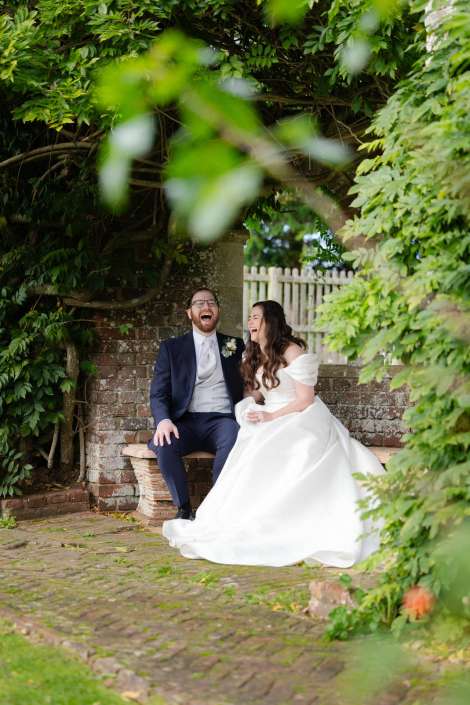 Bride in a white wedding dress and groom in a navy suit sit on a stone bench beneath a leafy arch, laughing together.