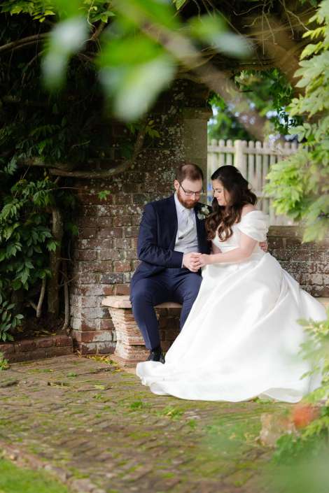 Bride in a white wedding dress and groom in a navy suit sit close together on a brick bench, holding hands beneath green vines.