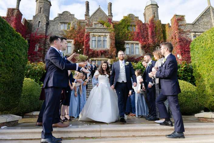 Bride in a white gown and groom in a suit walk down stone steps, flanked by bridesmaids in blue and groomsmen in navy, with an ivy-covered historic stone building in the background.