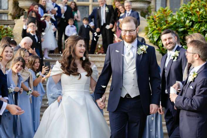 Bride and groom walk hand in hand down the steps as bridesmaids in blue dresses and groomsmen in navy suits cheer and toss petals.