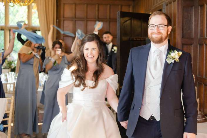 Happy bride in an off-the-shoulder white wedding dress and groom in a navy suit walk down the aisle as bridesmaids in blue celebrate in a wood-paneled room.