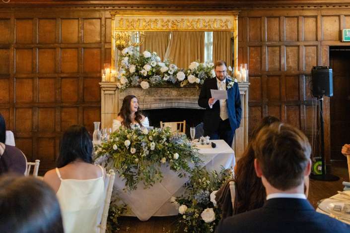 Bride at a flower-filled head table listening to a man in a suit giving a toast at a wedding in a wood-paneled room.