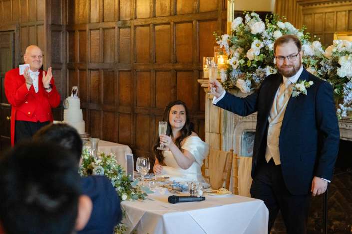 Bride in white and groom in a suit toast with champagne at a wood-paneled wedding reception, as a guest in a red jacket claps beside a large floral arrangement.