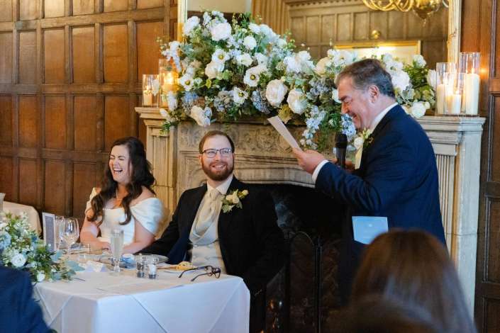 Bride and groom sit at a wedding reception table as a man in a suit gives a speech, with a large floral arrangement and candles behind them.