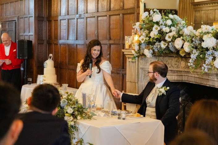 Bride in a white off‑the‑shoulder wedding dress speaks into a microphone while the groom sits beside her at a flower‑strewn head table, with a tall wedding cake and a wood‑paneled wall in the background.