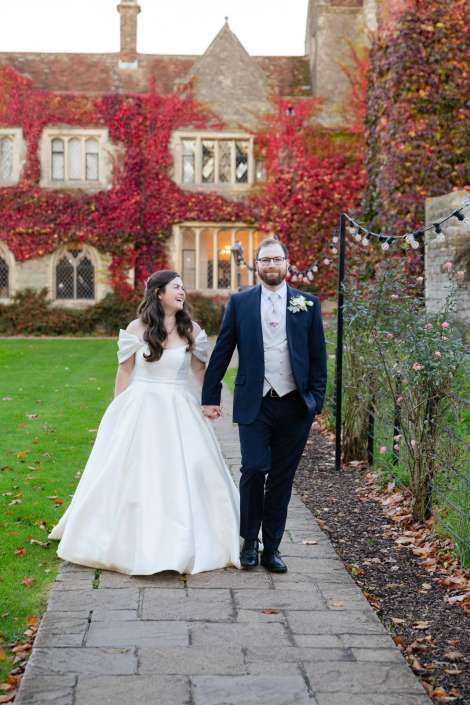 Bride in a white wedding gown and groom in a navy suit walk hand in hand along a stone path beside an ivy-covered historic building with red autumn leaves.