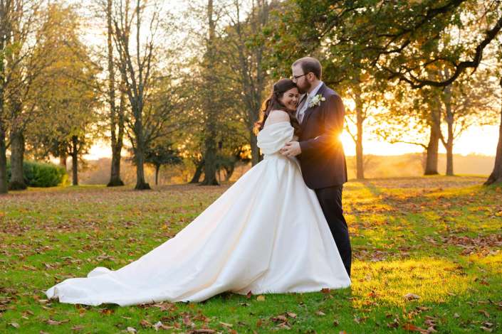 A bride and groom embrace in a sunlit autumn park, the bride in a long white wedding gown and the groom in a dark suit.