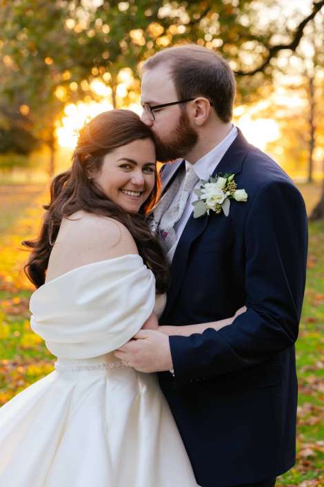 Bride and groom embrace outdoors at sunset, the groom kissing the bride on the forehead as she smiles.