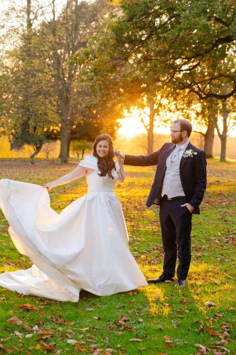 A bride in a white wedding gown twirls with the groom in a dark suit in a sunlit autumn park.