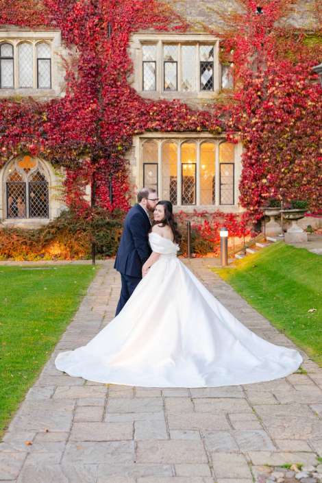 Bride in a white wedding gown with a long train and a groom in a dark suit pose on a stone path in front of an ivy-covered historic building with warm-lit windows.