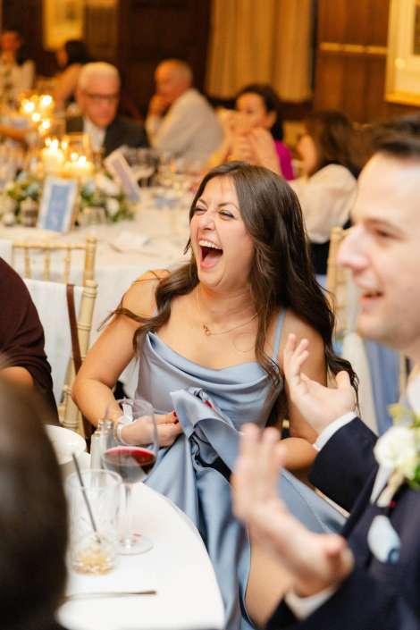 A woman in a light blue formal dress laughs joyfully at a wedding reception table with wine glasses.