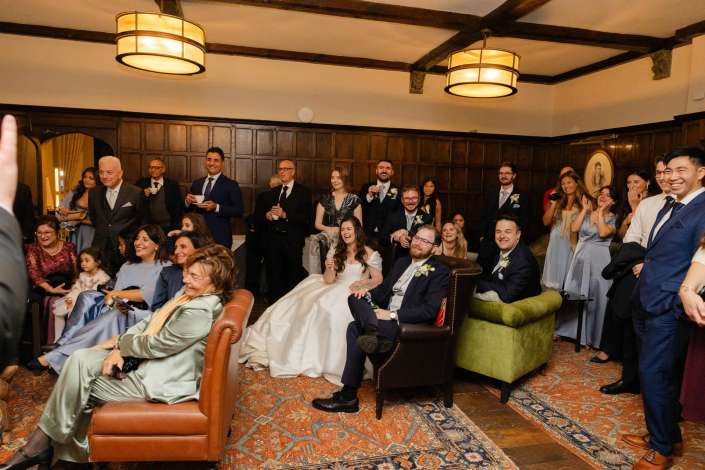 Guests in formal attire gather in a wood-paneled room for a wedding, with the bride in a white gown seated at center.