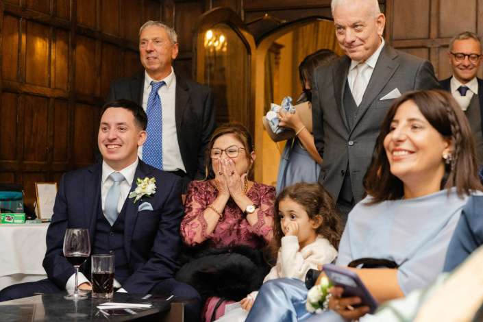 Formally dressed guests at a wedding reception indoors, seated around a table with wine, including a smiling man in a navy suit, a woman in burgundy, and a young child beside them.