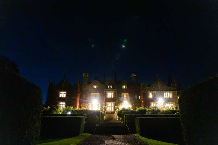 A grand stone mansion with warmly lit windows sits in a formal garden at night beneath a starry sky.
