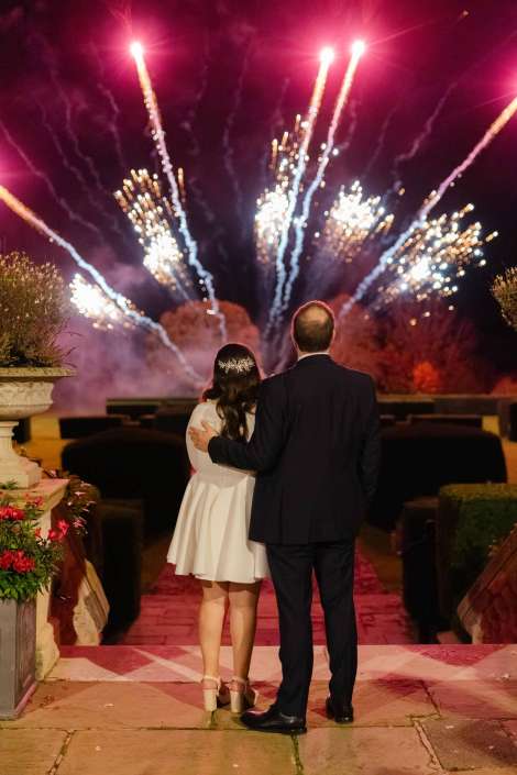 A couple stands with their backs to the camera, arm in arm, watching a fireworks display in the night.