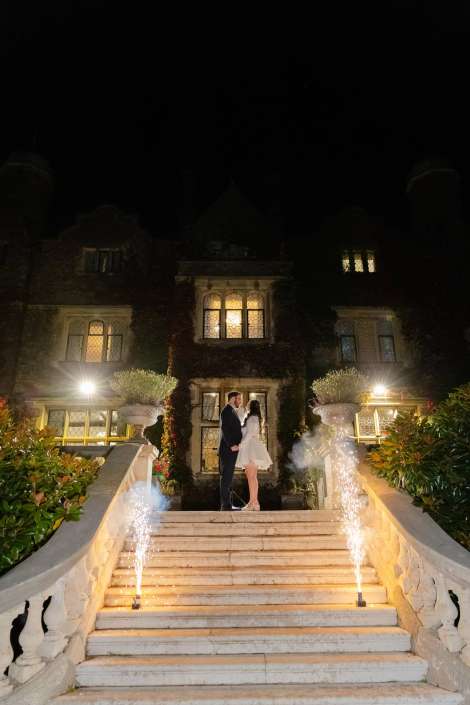 A bride and groom stand on illuminated marble stairs in front of an ivy-covered mansion at night, with sparkling fountains on either side.