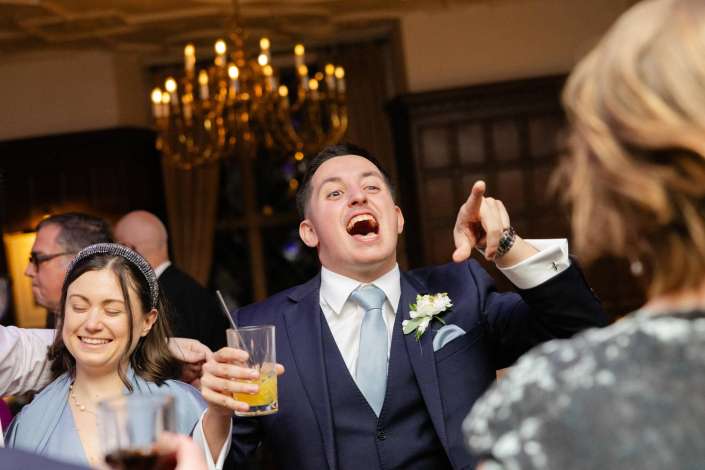 A man in a navy suit and light blue tie laughs and points while holding a drink at a wedding reception, with a smiling woman in blue beside him.