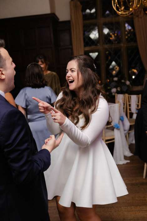A woman in a white dress laughs and points while dancing with a man in a dark suit at a wedding reception.