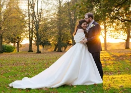 A bride and groom embrace in a sunlit autumn park, the bride in a long white wedding gown with a sweeping train.