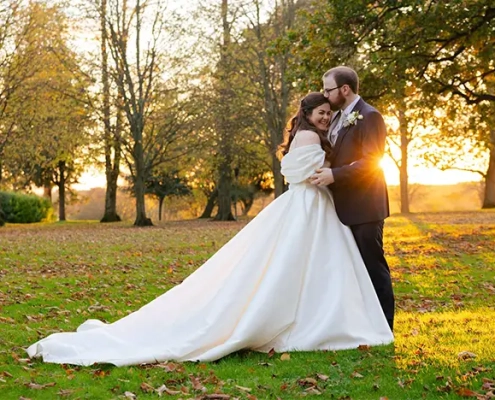 A bride and groom embrace in a sunlit autumn park, the bride in a long white wedding gown with a sweeping train.