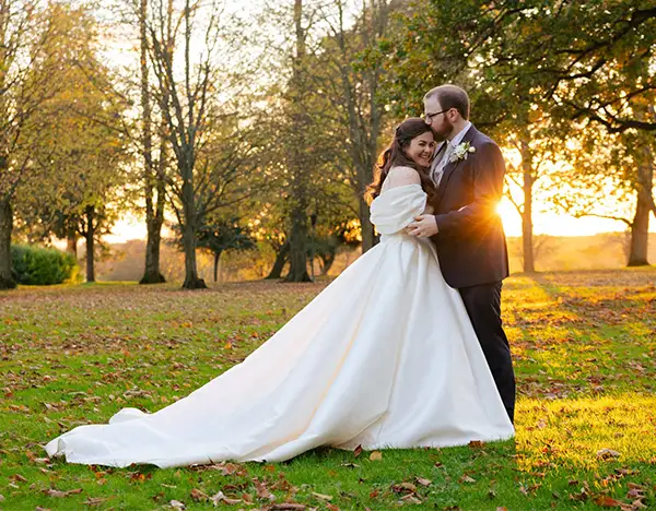 elizabetta & james, eastwell manor, louisa dettmer photography A bride and groom embrace in a sunlit autumn park, the bride in a long white wedding gown with a sweeping train.