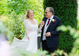 A bride in a white lace wedding gown walks hand in hand with a man in a dark suit through a lush garden, both smiling.
