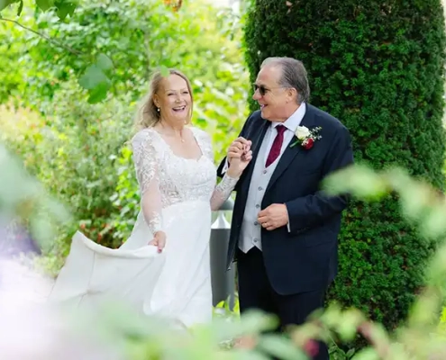 A bride in a white lace wedding gown walks hand in hand with a man in a dark suit through a lush garden, both smiling.