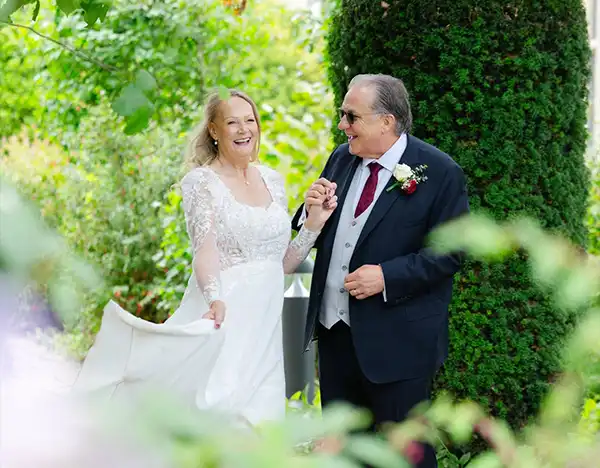 suzie & ian, eastwell manor, louisa dettmer photography A bride in a white lace wedding gown walks hand in hand with a man in a dark suit through a lush garden, both smiling.