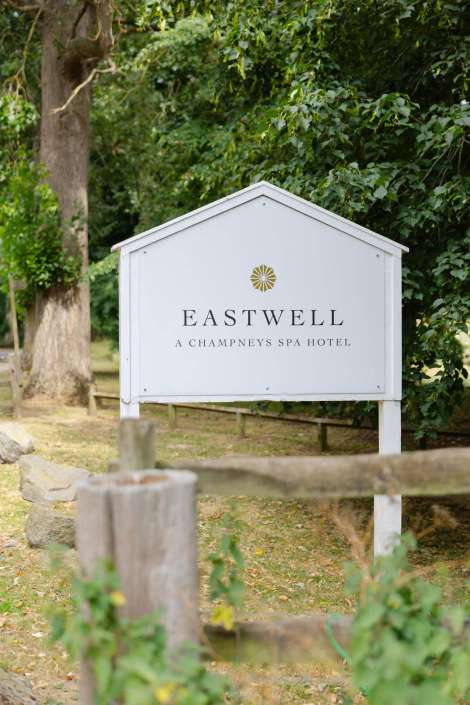 eastwell spa hotel sign A white signboard with a gold emblem and the text 'Eastwell' and 'A Champneys Spa Hotel' sits among green trees.