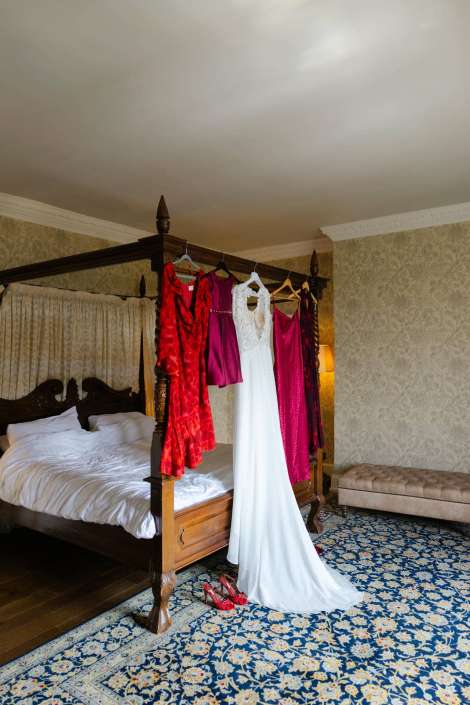 bridal gown and evening dresses White bridal gown and several colorful dresses hang from a four-poster bed in a warmly lit bedroom, with red heels on a blue floral rug.