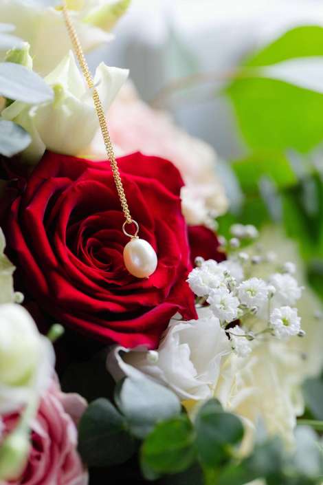 pearl necklace on red rose A red rose in a bouquet with white blooms and greenery, with a gold chain and pearl pendant draped over its petals.