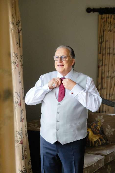 gentleman adjusting his tie Smiling elderly man with gray hair and glasses, wearing a light gray vest and white shirt, adjusting a red tie indoors near a window.