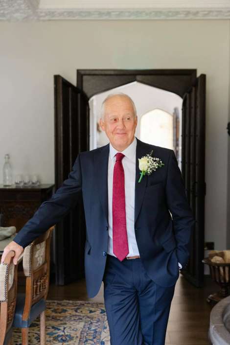 man in navy suit with boutonniere An elderly man in a navy suit with a red tie and a white boutonniere stands in a doorway indoors, smiling.