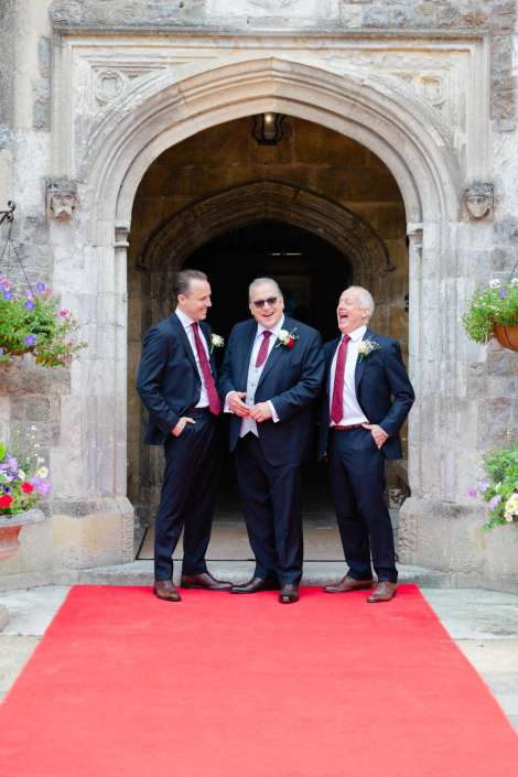 groomsmen on red carpet Three men in dark suits with red ties stand on a red carpet outside a stone arched doorway, smiling and chatting.