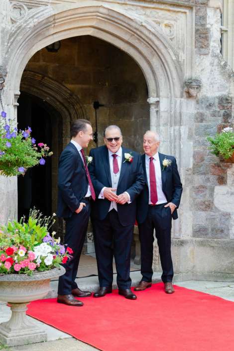 groomsmen at church entrance Three men in dark suits with red ties and white boutonnieres stand on a red carpet beneath a stone archway, chatting beside flowering planters.