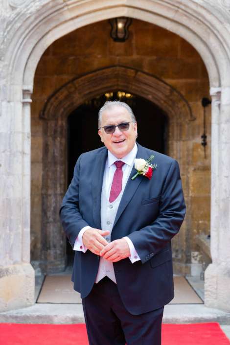 groom at church entrance Smiling man in a dark suit with a boutonniere and sunglasses stands on a red carpet in front of a stone arched doorway.