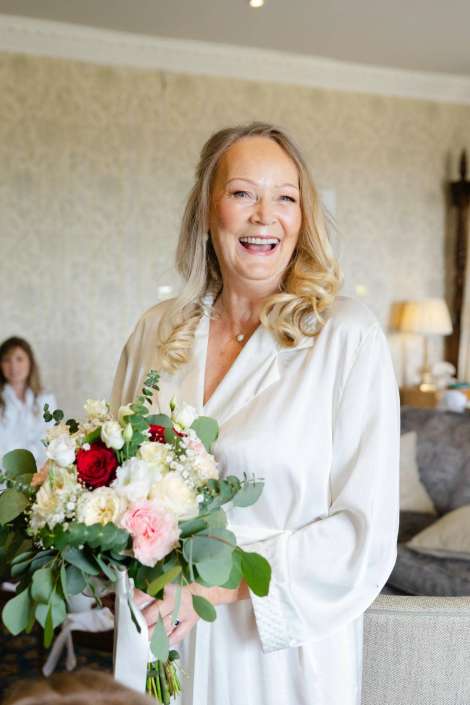 joyful bride with bouquet Smiling woman in a white robe holding a bouquet of pink, red, and white flowers with greenery indoors.