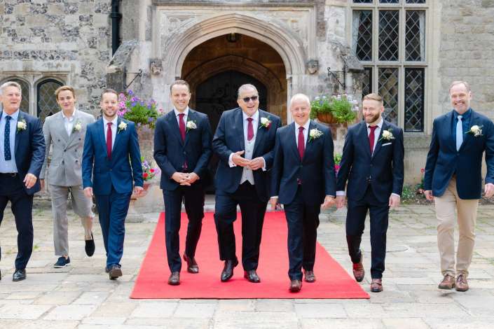 groomsmen arrive on red carpet Group of men in suits walking on a red carpet outside a historic stone building, wearing boutonnieres.