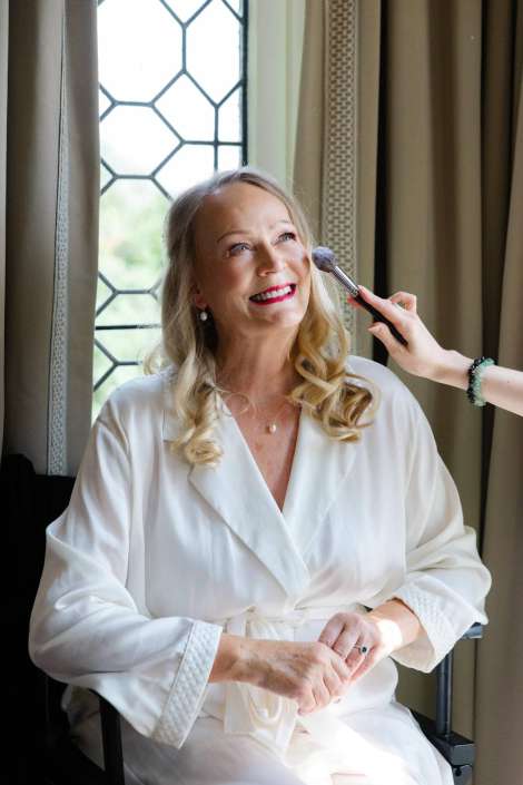 radiant bride getting ready Smiling blonde woman in a white robe sits by a window while makeup is applied with a brush.