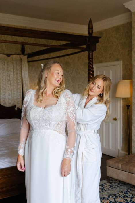 bride getting ready with friend Bride in a white lace wedding dress stands in a bedroom as another woman in a white robe helps her with her hair.