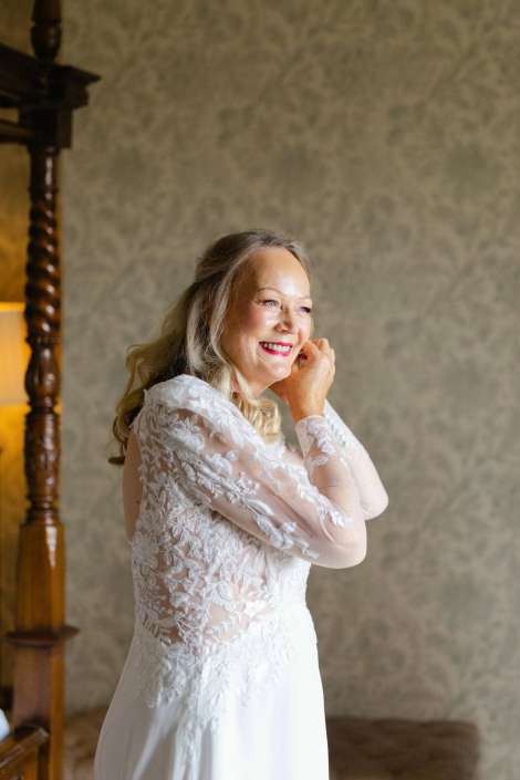 smiling bride in lace dress A smiling older woman in a white lace wedding dress stands indoors, gently touching her hair.