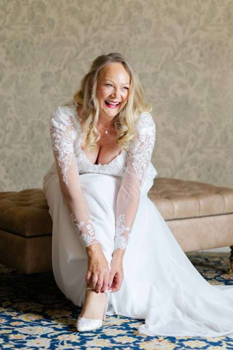 joyful bride in lace dress Smiling bride in a lace wedding gown sits on a tufted bench and puts on her white high-heeled shoes.