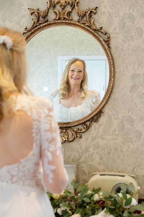 bride reflected in mirror Smiling bride in a white lace wedding dress looks at her reflection in an ornate round mirror on a patterned wall.