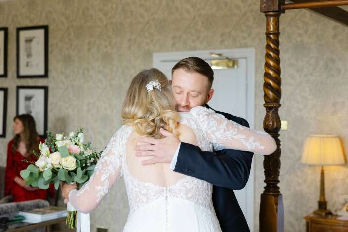 bride and groom hug Bride in a lace wedding dress hugs a man in a dark suit indoors, holding a bouquet of flowers.