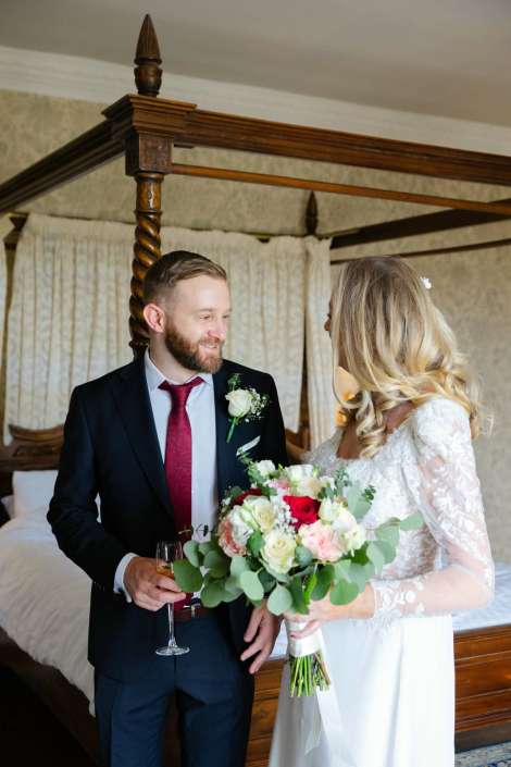 bride and groom in canopy bed Bride in a lace wedding dress and a groom in a dark suit with a red tie smile at each other indoors beside a carved four-poster bed, as the bride holds a bouquet of white, pink, and red flowers with greenery.