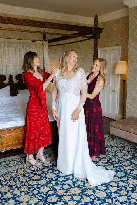 bride surrounded by bridesmaids A bride in a white lace wedding gown stands between two bridesmaids in red dresses, as they adjust her hair in a bedroom with a four-poster bed.