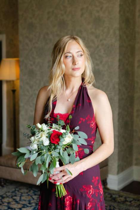 blonde bride with bouquet A blonde woman in a burgundy floral halter dress holds a bouquet of white and red flowers with greenery indoors.