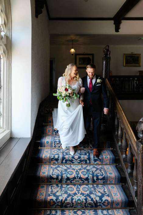 bride and groom descending staircase Bride in a white lace wedding dress carries a bouquet as she walks down a patterned staircase with a groom in a dark suit beside her.
