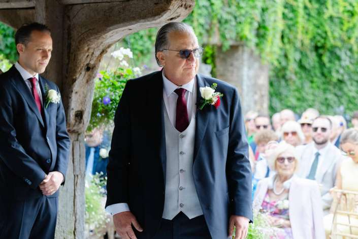 elegant gentleman at garden wedding An older man in a dark suit and gray waistcoat with a white boutonniere and sunglasses stands at an outdoor wedding.