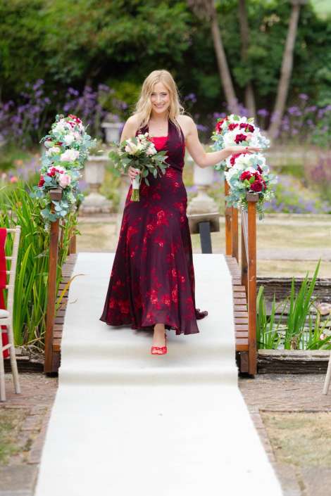 bride walking down garden aisle Smiling woman in a burgundy floral gown walks down a white aisle on a garden bridge, holding a bouquet and flanked by flower arrangements.